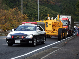 写真：【警察のパトロールカーの先導により、道路維持作業車両（グレーダー）で走行不能のトレーラーを移動】