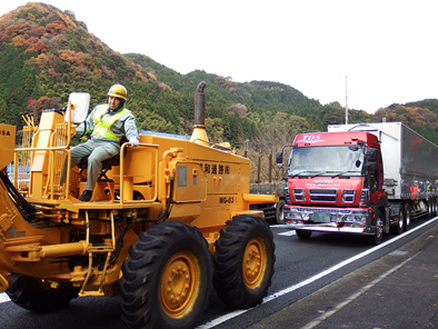 写真：【警察のパトロールカーの先導により、道路維持作業車両（グレーダー）で走行不能のトレーラーを移動】